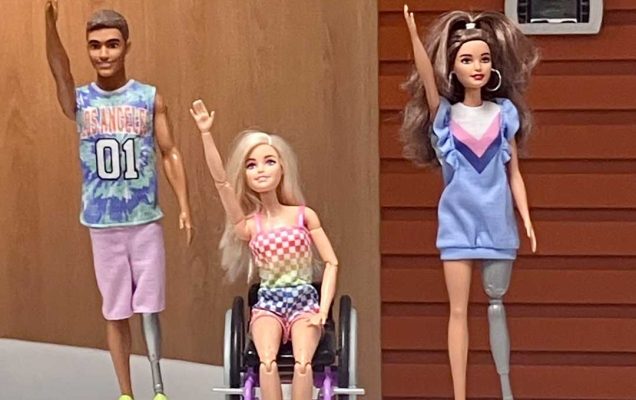 Dwie Figurki Barbie i Kena stojących obok siebie. Ken i Barbie stojący po bokach mają protezy nóg, między nimi siedząca na wózku Barbie. Wszystkie postaci mają podniesioną do góry prawą rękę.