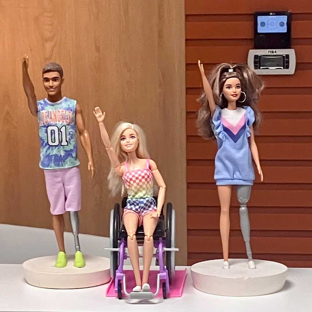 Dwie Figurki Barbie i Kena stojących obok siebie. Ken i Barbie stojący po bokach mają protezy nóg, między nimi siedząca na wózku Barbie. Wszystkie postaci mają podniesioną do góry prawą rękę.