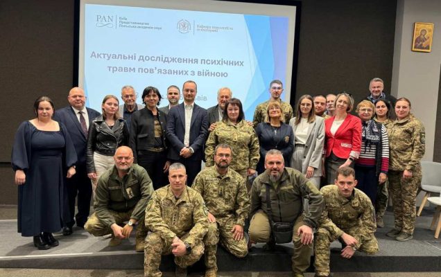 Zdjęcie grupowe uczestników w strojach cywilnych i wojskowych na sali konferencyjnej. Za nimi wielki ekran z wyświetlonym w języku ukraińskim napisem prezentacji.