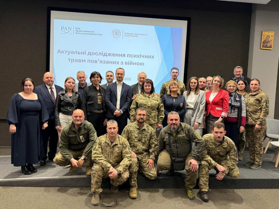 Zdjęcie grupowe uczestników w strojach cywilnych i wojskowych na sali konferencyjnej. Za nimi wielki ekran z wyświetlonym w języku ukraińskim napisem prezentacji.