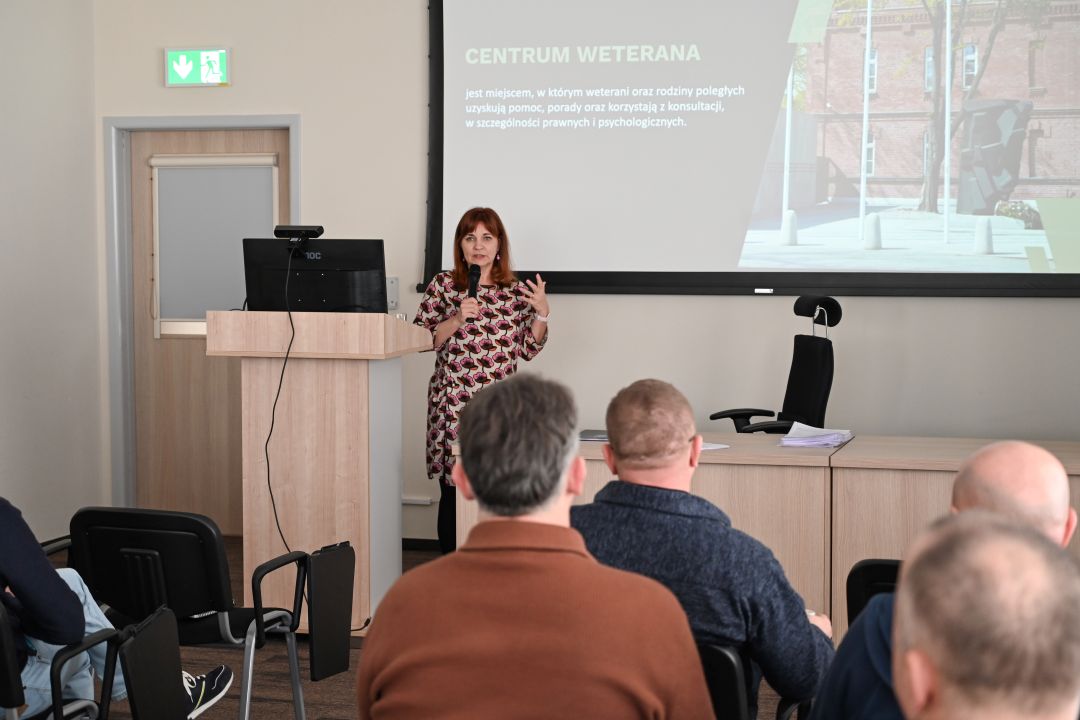 Siedzący słuchacze podczas wykładu. Prelegentka prowadzi wykład. Za jej plecami na dużym ekranie plansza prezentacji.