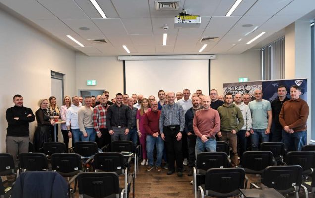 Zdjęcie grupowe stojących uczestników wydarzenia na sali wykładowej.