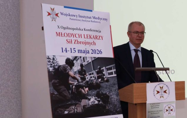 Prelegent przy mównicy, obok niego na stojaku plakat konferencji.