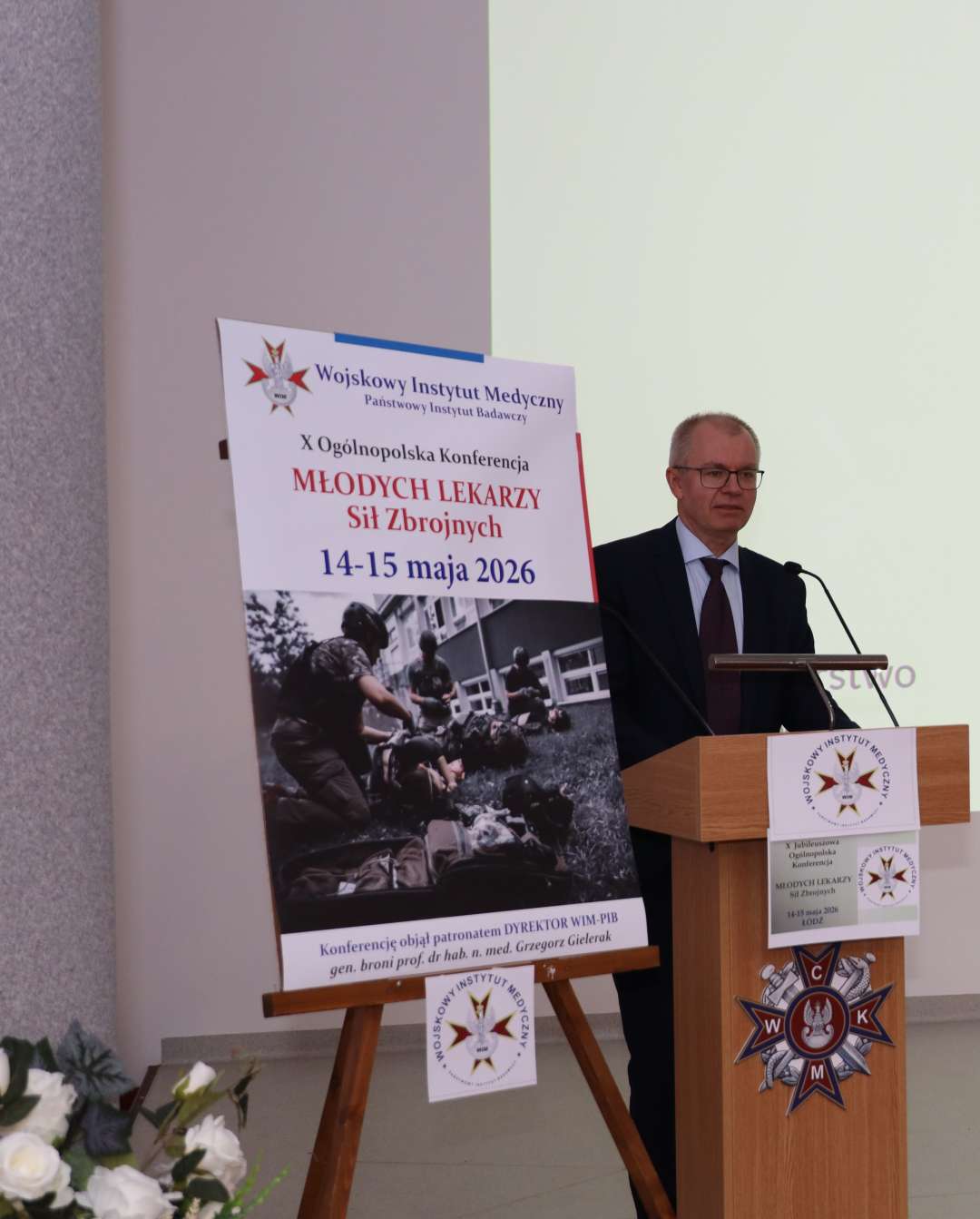 Prelegent przy mównicy, obok niego na stojaku plakat konferencji.