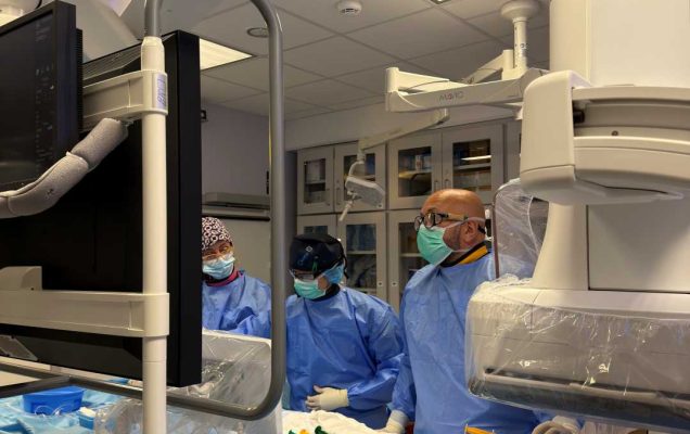 Trzech lekarzy w sali zabiegowej wykonuje procedurę medyczną przy pacjencie przykrytym niebieskim sterylnym materiałem; personel w maskach i fartuchach operacyjnych pracuje przy stole z aparaturą i monitorami medycznymi w nowoczesnym pomieszczeniu szpitalnym.