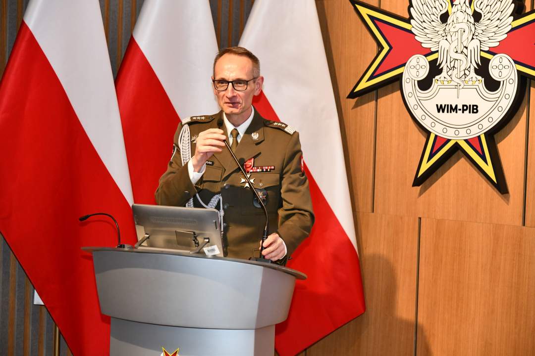 Grzegorz Gielerak przy mównicy. Za nim trzy flagi Polski i logotyp WIM.