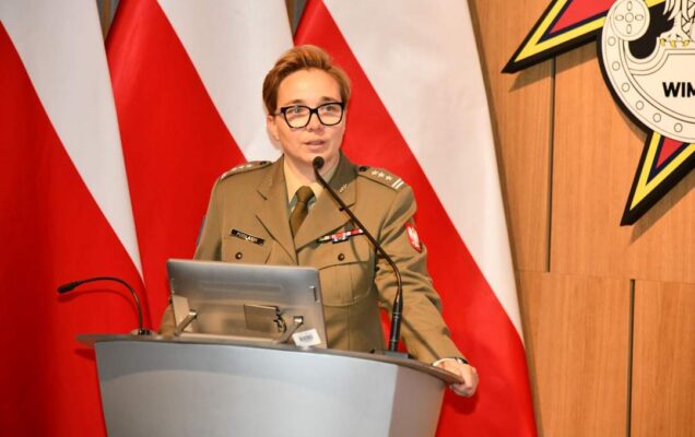 płk Anita Podlasin - Dyrektor Departamentu Wojskowej Służby Zdrowia przy mównicy. Za nim trzy flagi Polski i logotyp WIM.