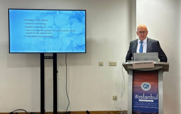 Prelegent przy mównicy podczas prelekcji. Obok niego na ekranie wyświetlony slajd z konferencji.