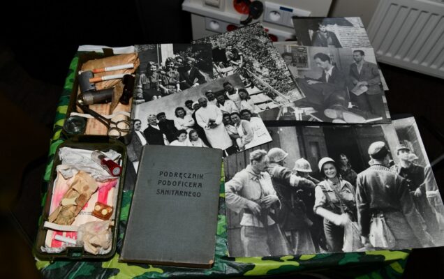 Stara apteczka polowa, podręcznik podoficera sanitarnego oraz czarno-białe fotografie leżące na stole.