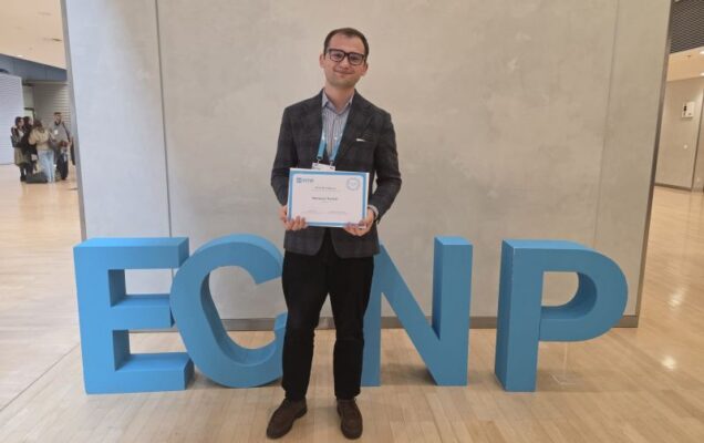 Mateusz Kozioł z dyplomem w holu konferencji. Za jego plecami instalacja artystyczna z logo konferencji ECNP.