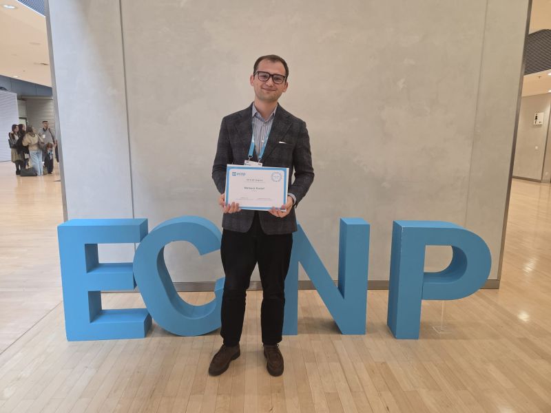 Mateusz Kozioł z dyplomem w holu konferencji. Za jego plecami instalacja artystyczna z logo konferencji ECNP.